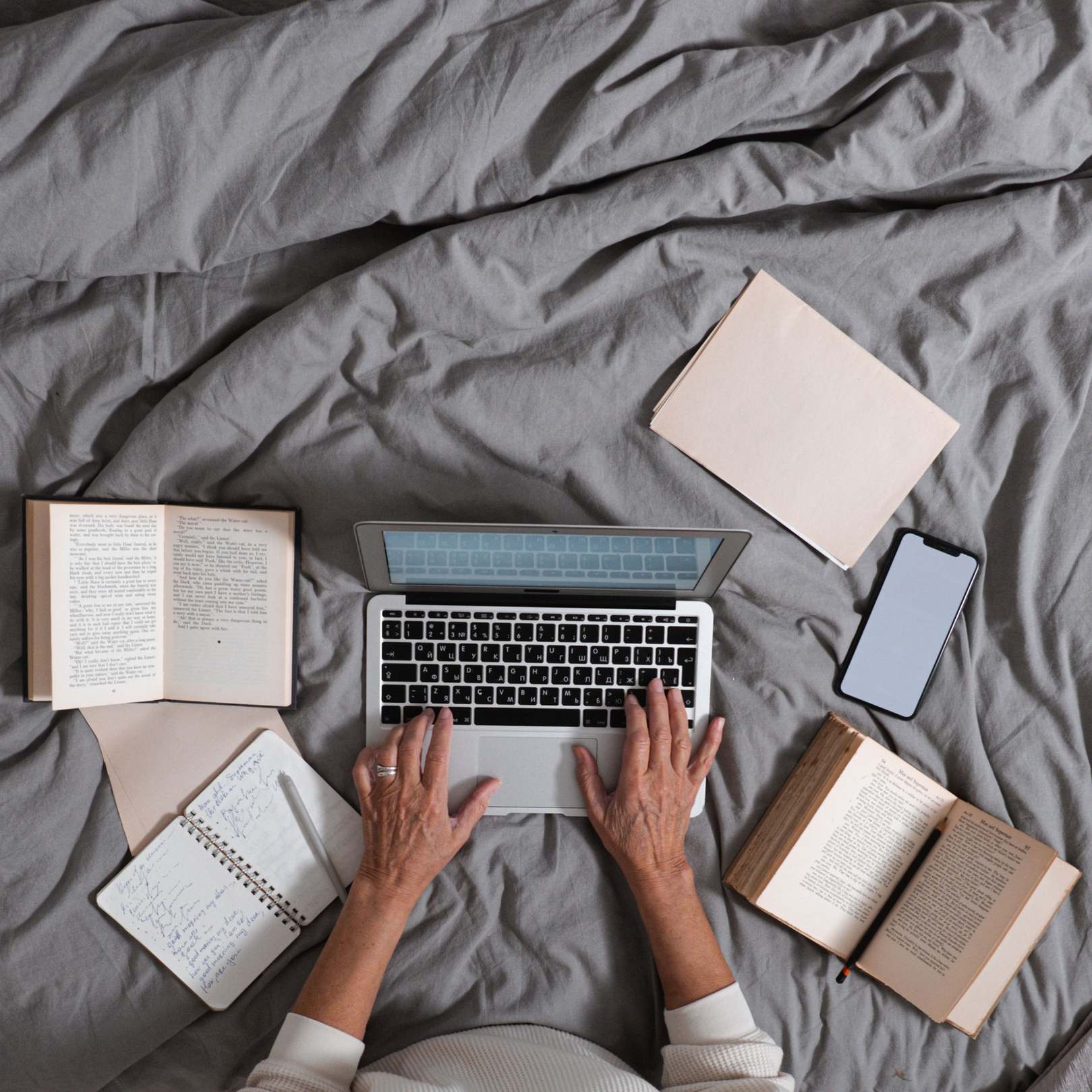 Overhead view of a laptop workspace — person typing on laptop surrounded by books and smartphone on a bed, top view